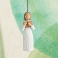 Willow Tree Love of Learning Ornament, 4”h hand-painted resin figure in cream dress holding open book to her chest, brown elastic cord, hanging in front of cyan painting