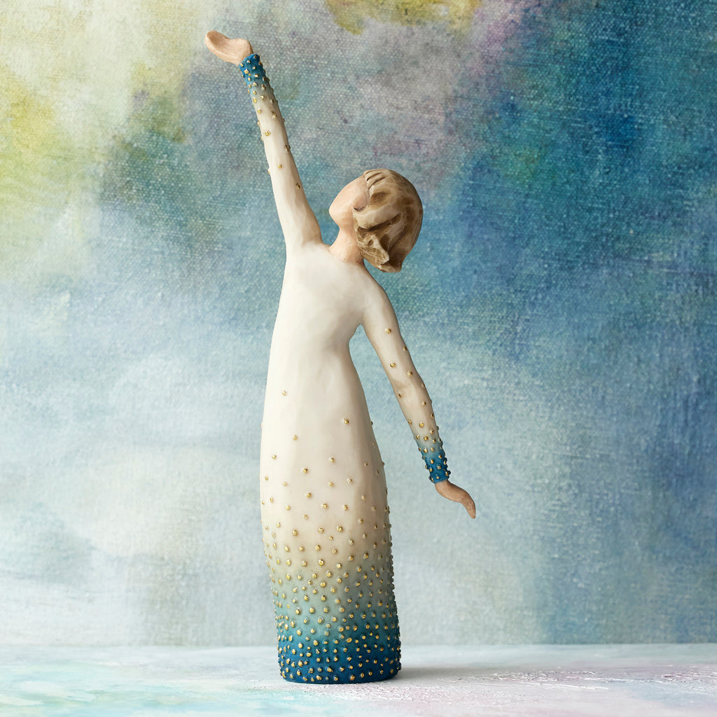 Willow Tree Shine, 8" hand-painted resin figure in cream and blue ombré dress with gold-leaf dots, looking up, one arm extended upward, in front of ombré blue painting