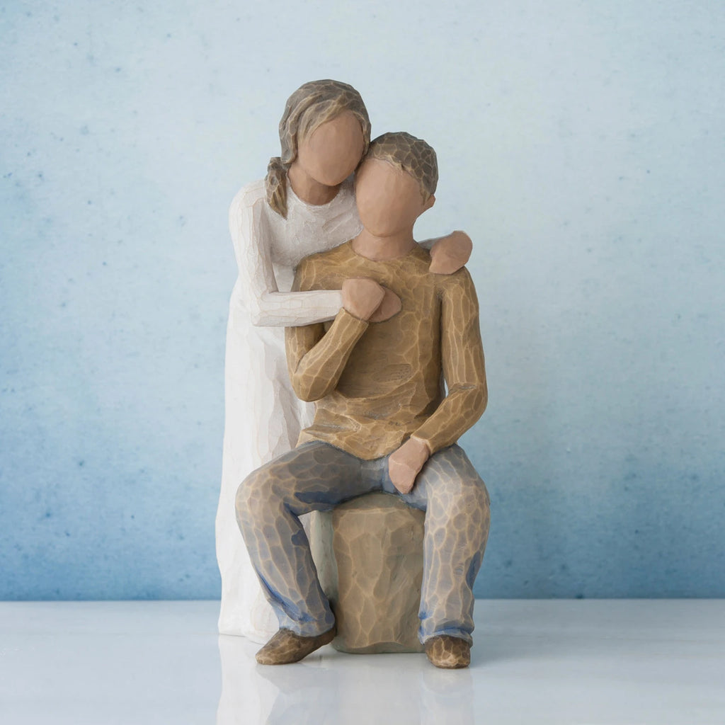 Man figurine sitting on rock wearing brown shirt and blue jeans with woman figurine in white dress with arms wrapped around him - blue background