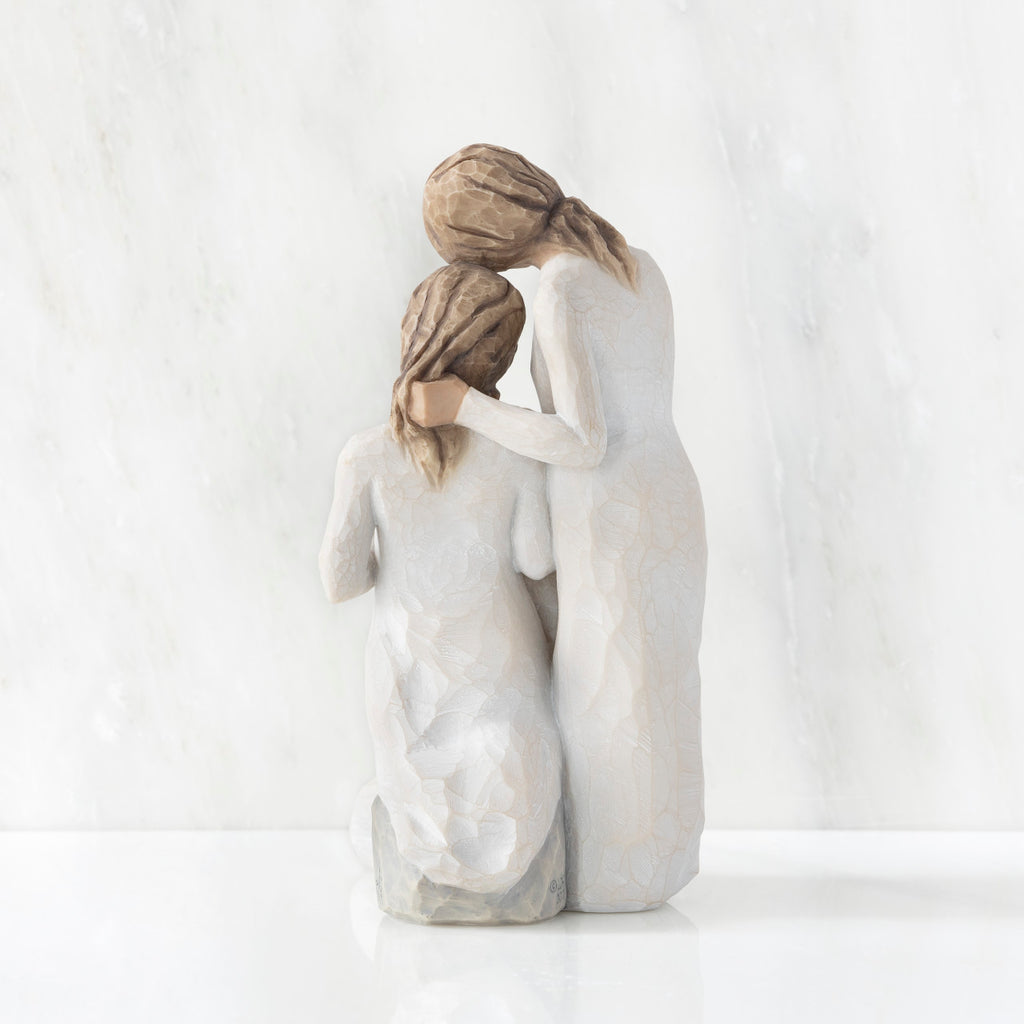 Back view, figurine of three females in cream dresses: one standing with arm around kneeling female, and holding hand of seated female