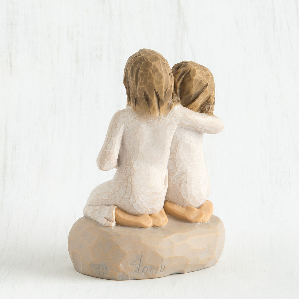 Back view of two young brunette girls in white sitting on a tan rock with arms around each other