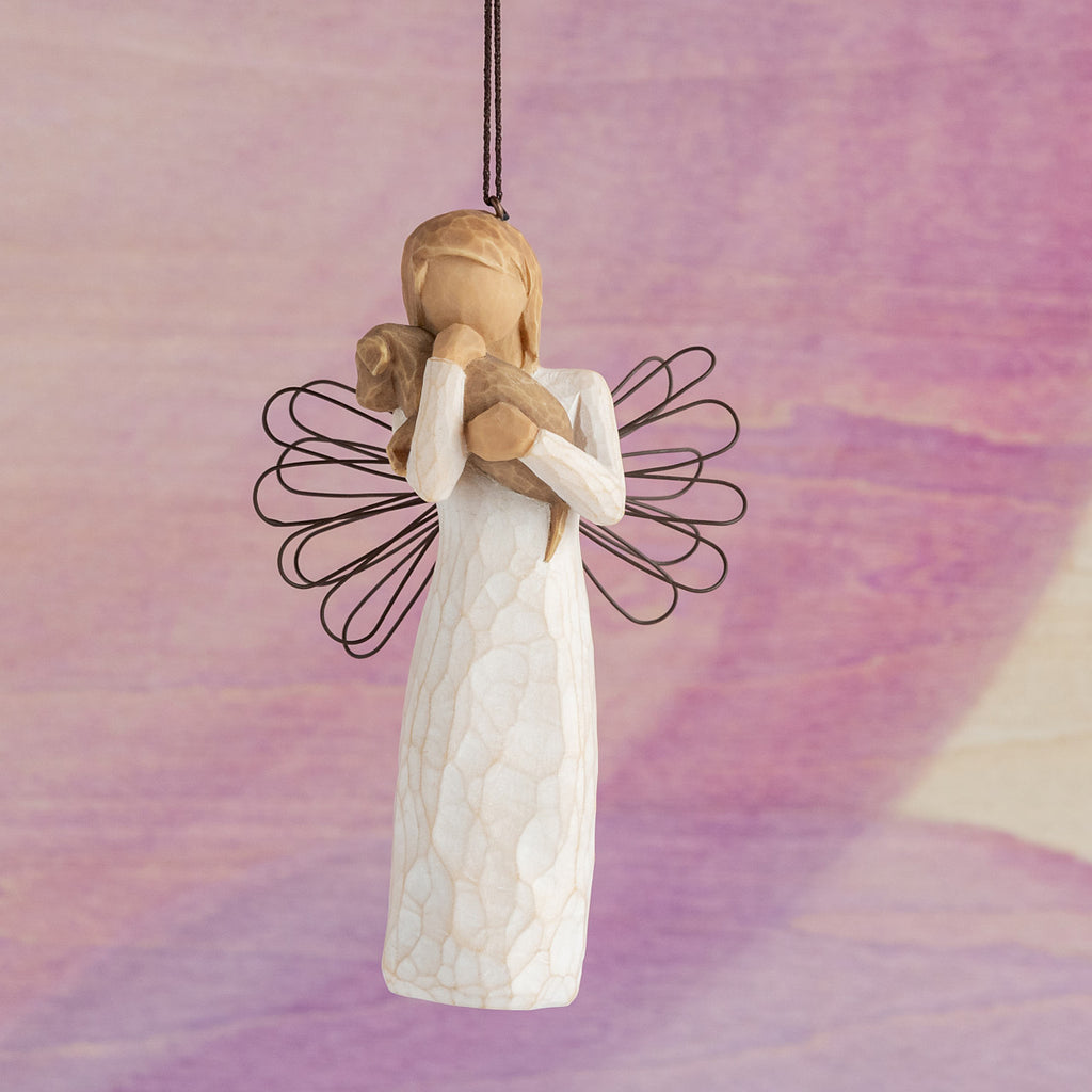Willow Tree Angel of Friendship Ornament, 4.5"h hand-painted resin figure in cream dress with wire wings holding brown dog, brown elastic cord, hanging in front of pink painting