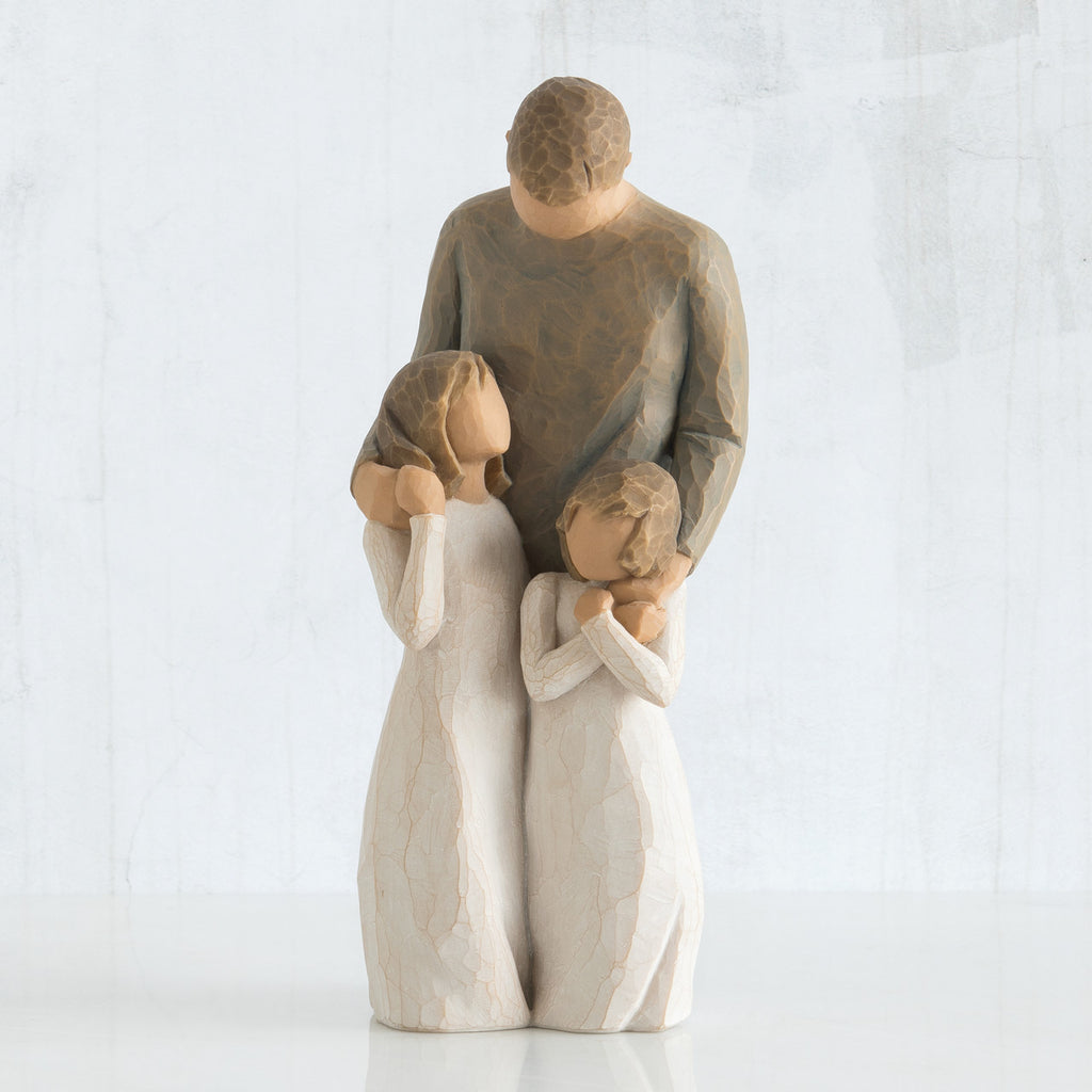 Man faceless figurine wearing brown shirt standing over two faceless girl figurines in white dresses
