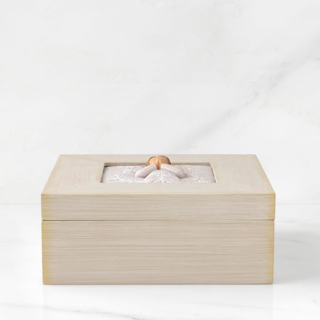 Side View: Cream square wood box with hinged lid, carving on lid of female in cream dress with hands in prayer position, surrounded by leaves and vines