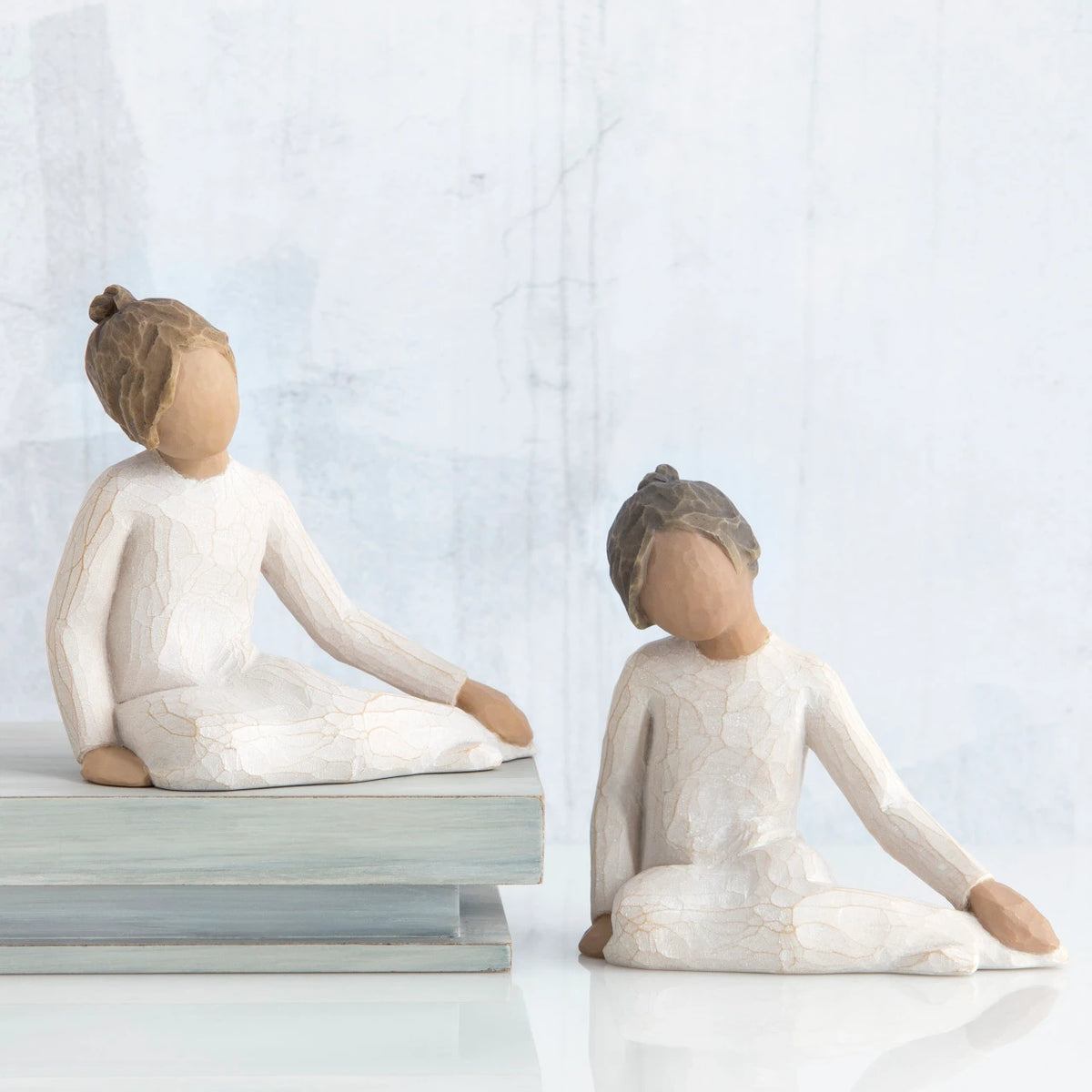 Two young brunette girl figurines in white sitting down next to wooden stack of books