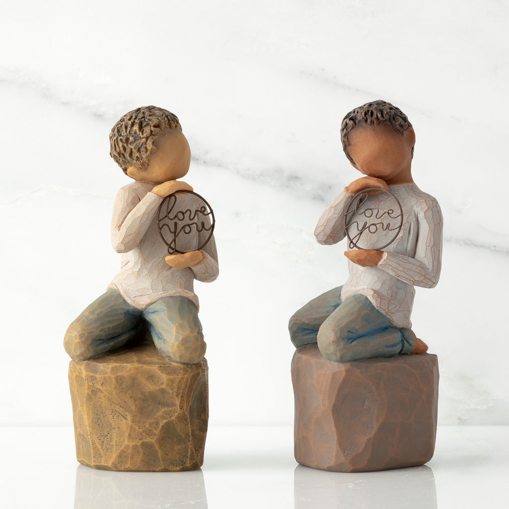 Two options (lighter skin and darker skin) of boy figurine kneeling on rock, holding wire circle with ‘love you’ inside