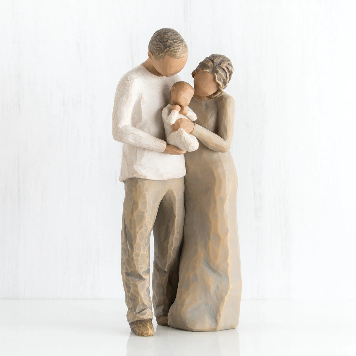 Man and woman faceless figurines holding baby in white outfit - man is dressed in white shirt and brown pants, woman is dressed in brown dress