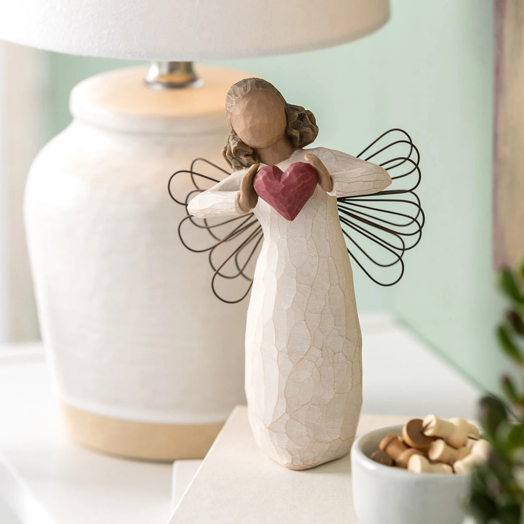 Willow Tree With Love, 5.5”h hand-painted standing resin angel figure with darker skin and hair, in cream dress with wire wings, holding a red heart in hands, on shelf with cream lamp