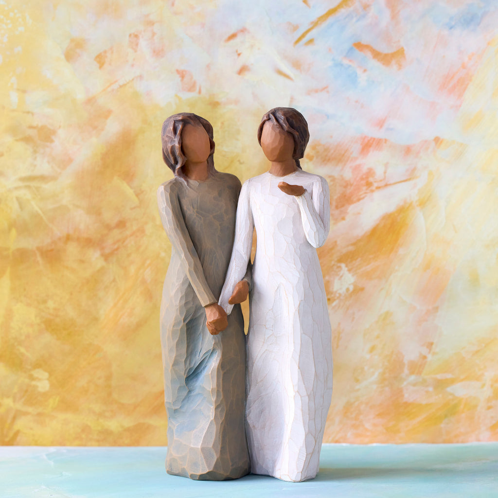 Willow Tree My sister, my friend, 8.5”h hand-painted standing figure of two women with darker skin and hair, standing arm in arm, one in cream dress, one in blue dress, in front of orange painting