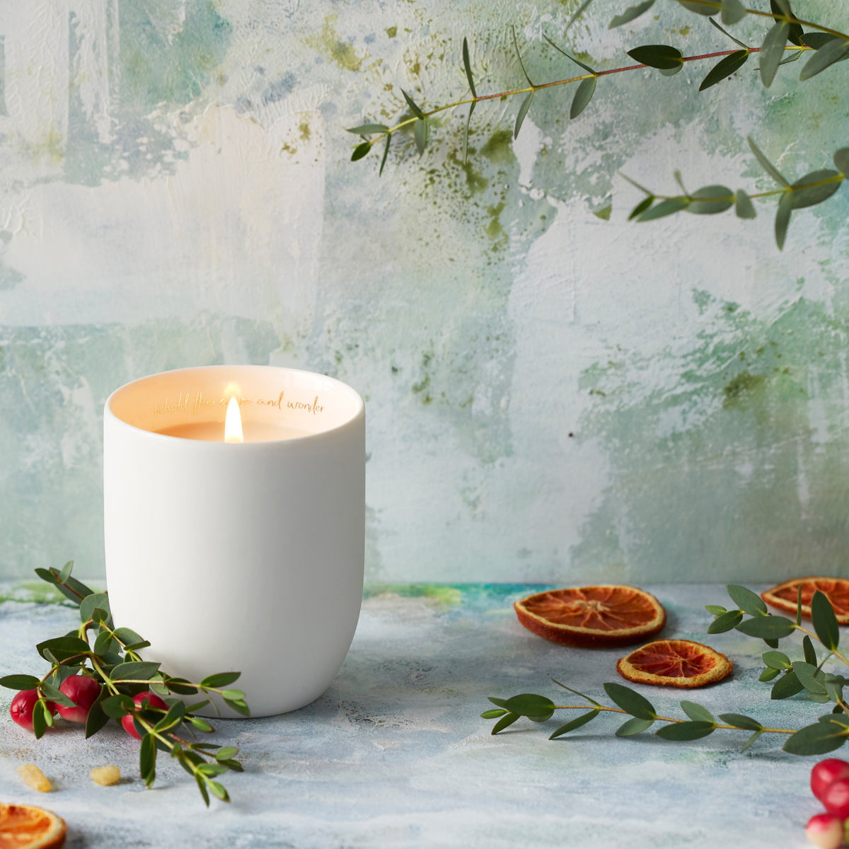 Willow Tree Festive Orange and Frankincense Candle, 3.5"h off-white ceramic vessel with plant-based wax and blend of natural oils, "behold the awe and wonder" sentiment in gold inside rim, in front of blue painting