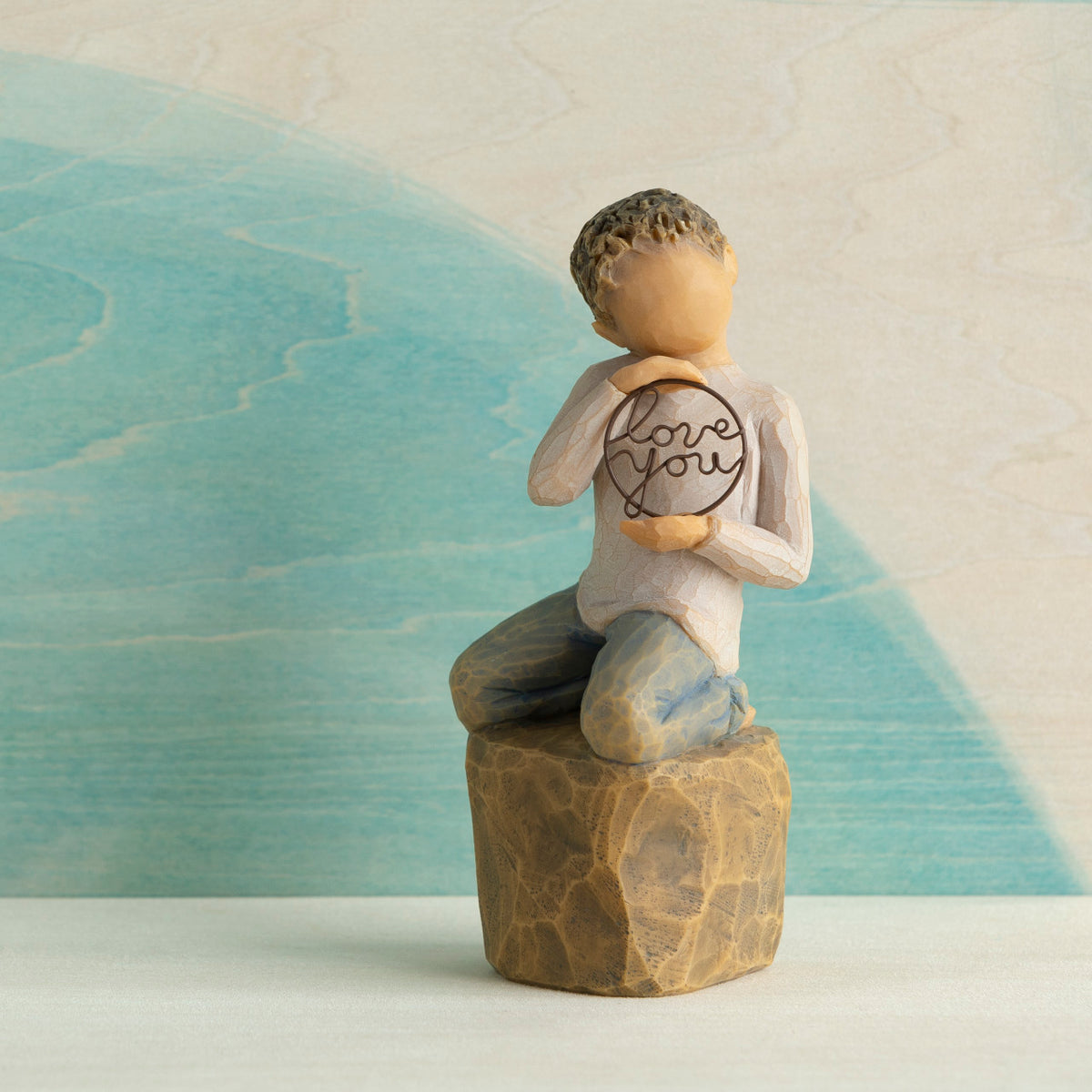 Willow Tree Love you too (lighter skin), 4.5”h hand-painted resin figure of young child in cream shirt and jeans, kneeling on rock, holding wire circle with ‘love you’ inside, in front of blue painting