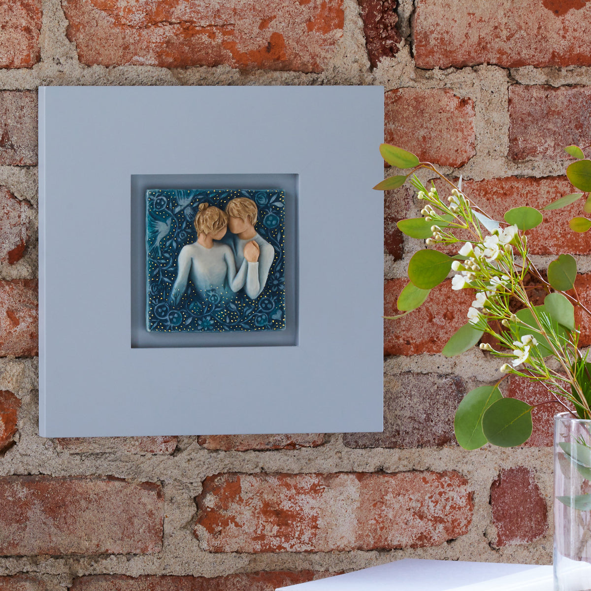 Willow Tree Duet, 10”sq hand-painted dove gray wood frame, with inset bas-relief resin plaque of embracing couple, surrounded by nature, blue ombré background with gold leaf, hanging on brick wall