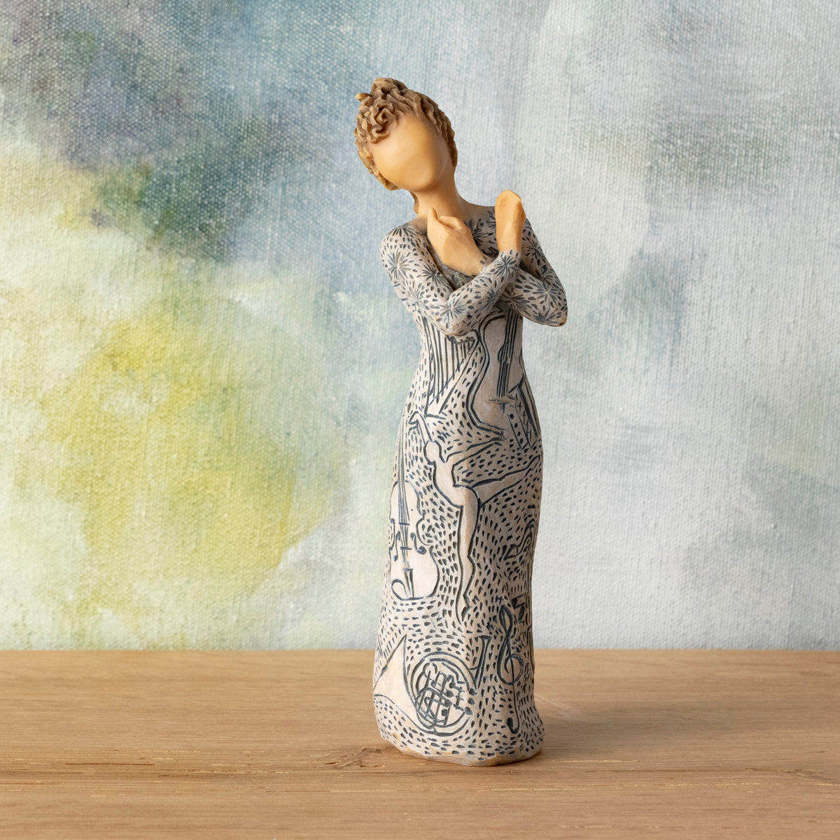 Willow Tree Music Speaks (lighter skin), 7”h hand-painted standing resin figure, arms crossed, wearing dress carved with symbols of music and dancing, in front of blue-green painting
